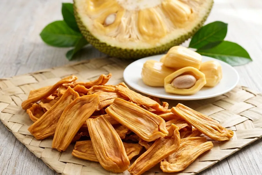 Dried jackfruit pieces on a woven palm mat in front of fresh jackfruit bulbs on a white plate and a cut jackfruit