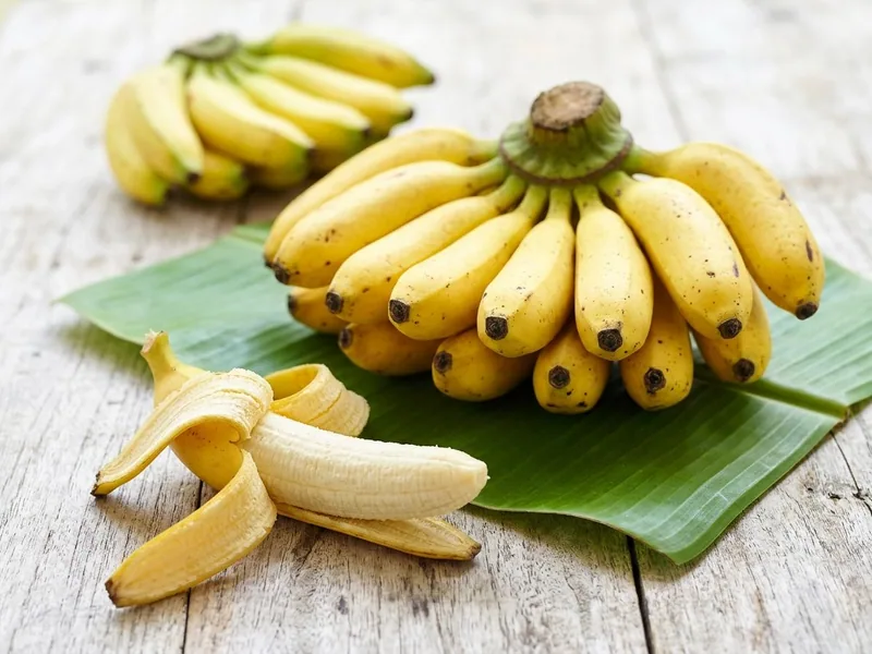 Hand of ripe bananas on a banana leaf, one banana half peeled, on a pale timber table