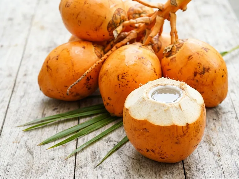 Orange king coconuts on their bunch stalk, one cut open at the top to the coconut water, on a pale timber table