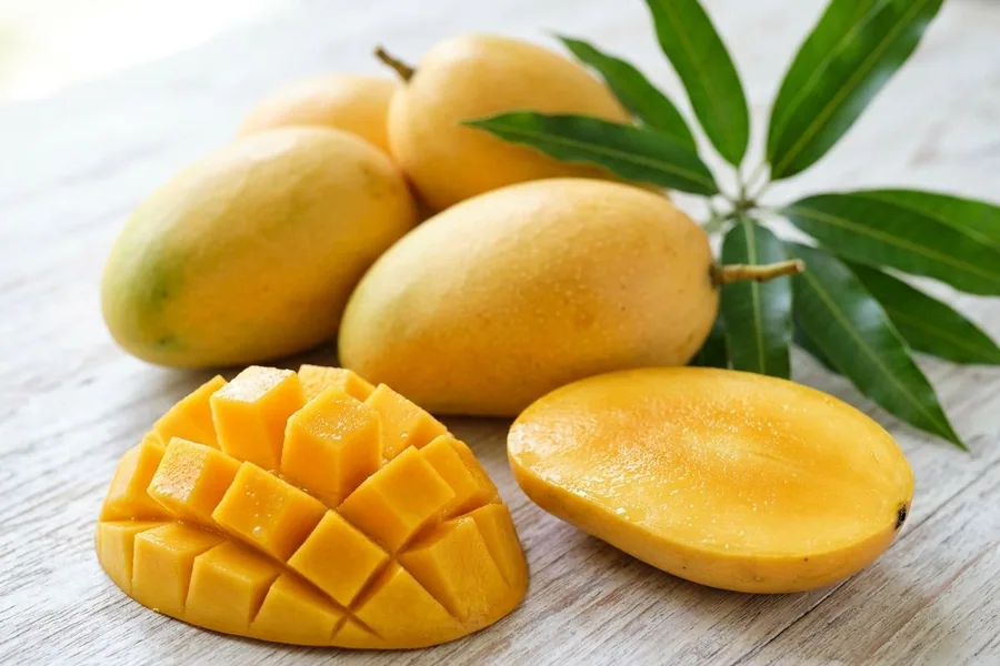 Ripe golden mangoes on a pale timber table, one cheek cut into cubes and one halved, with a sprig of mango leaves