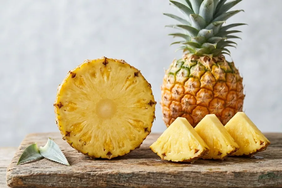Pineapple slice and three wedges in front of a whole pineapple on a rustic timber board