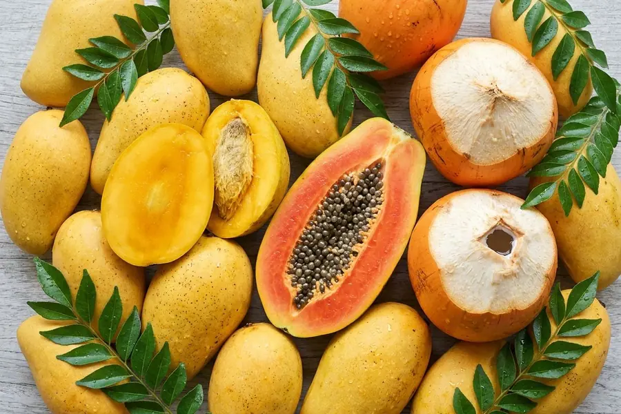 Ripe TJC mangoes, a Red Lady papaya halved to its seeds and orange king coconuts cut open at the top, with sprigs of curry leaves on pale timber