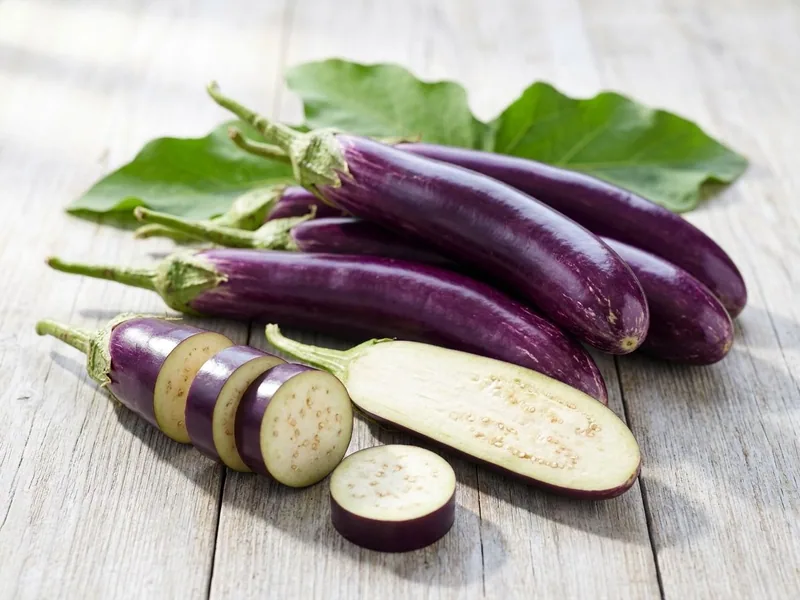 Long purple brinjals on a pale timber table, one sliced into rounds and one halved lengthways to the cream flesh
