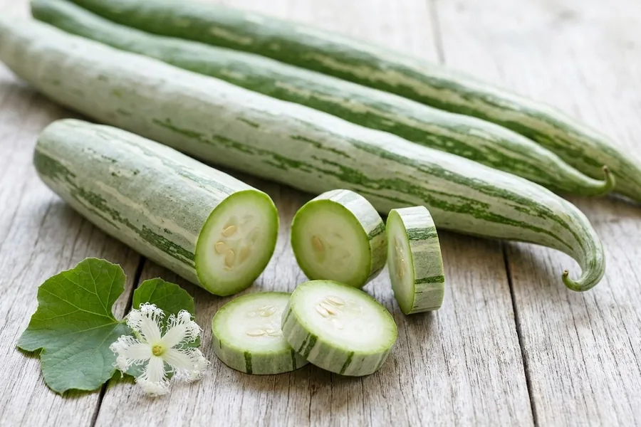 Long striped snake gourds on a pale timber table, one cut into rounds, with a snake gourd leaf and its fringed white flower