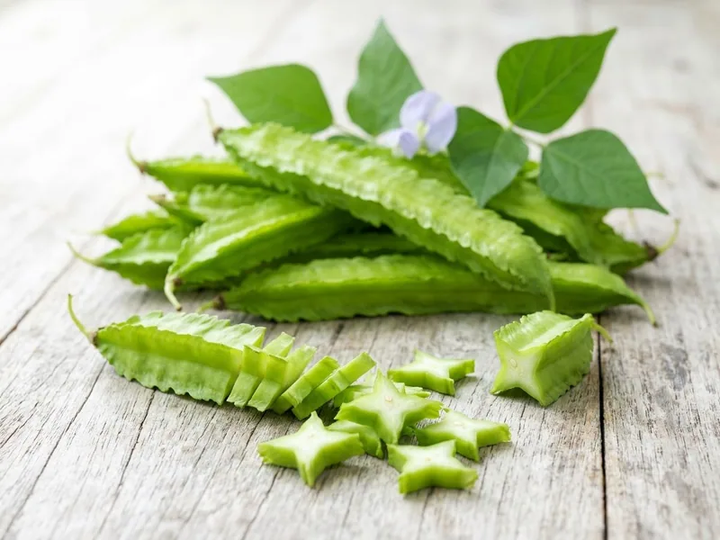 Winged bean pods with frilly edges on a pale timber table, one sliced into star-shaped pieces, with leaves and a pale blue flower