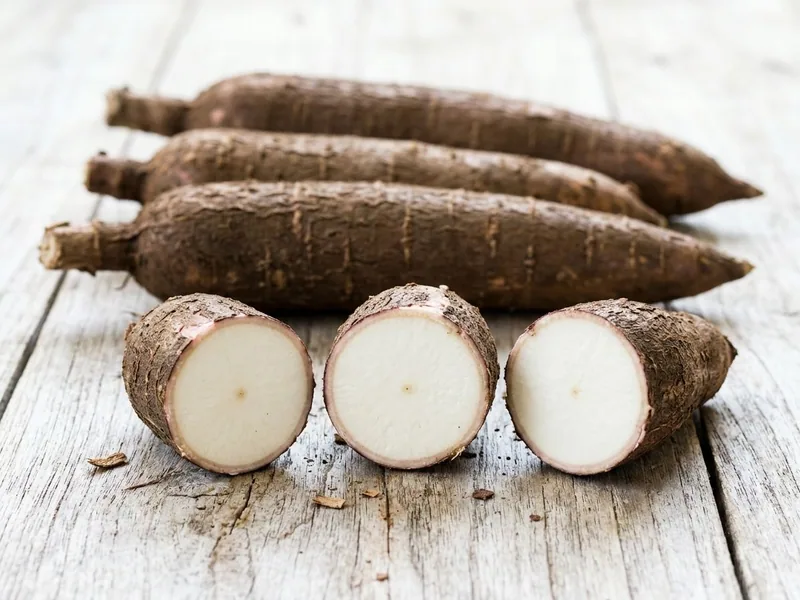 Cassava roots on a pale timber table, one cut into thick rounds showing white flesh inside the pinkish inner peel