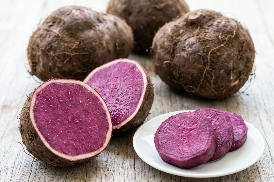 Whole purple yams on a pale timber table, one cut in half to its violet flesh, with slices on a small white plate