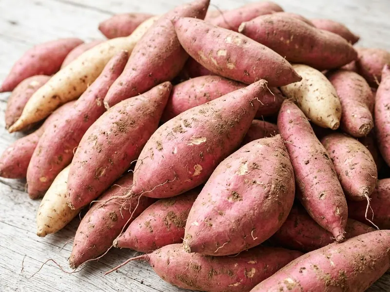 Whole sweet potatoes heaped on a pale timber table, most with reddish-pink skin and a few pale cream, with traces of soil and fine roots